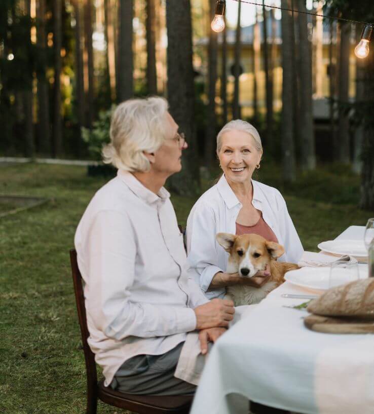 Older couple dressed in white sitting down for a white-table-cloth dinner, outside in lush greenery, with their Corgi